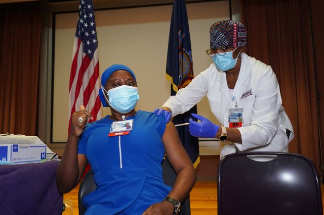Gemma John, in a mask, gets the vaccine from another nurse in a mask. They are both Black women.